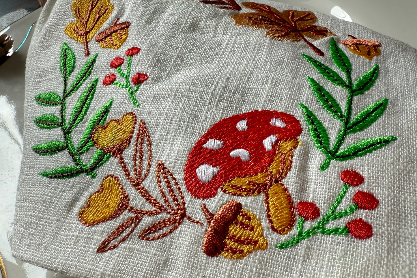 Embroidered fabric with red mushroom, green leaves, and acorns on a white background