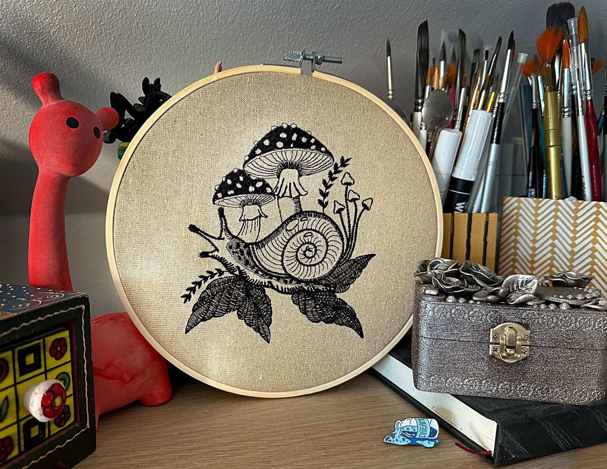 Embroidery hoop with black and white snail and mushroom design on a wooden surface with art supplies.