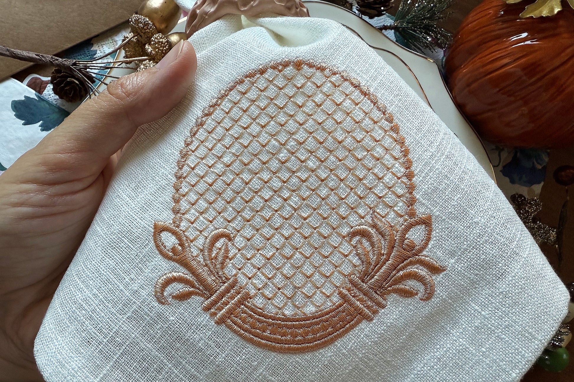 Embroidered design on a white fabric held by a hand, with a blurred natural background.