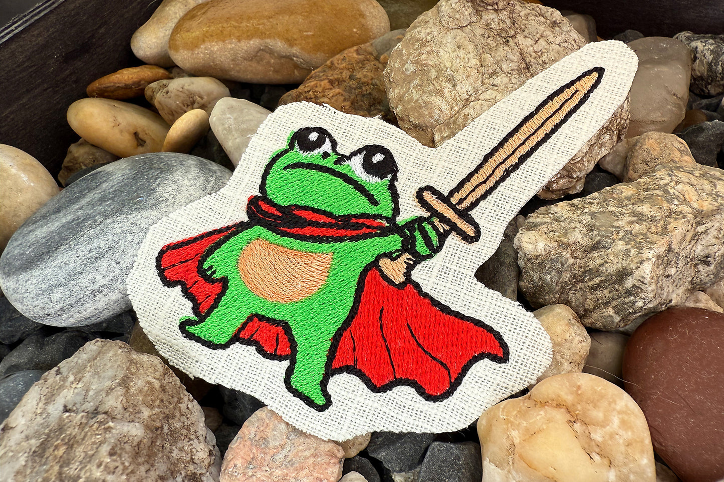 Embroidered patch of a frog in a superhero costume with a sword on a bed of stones.