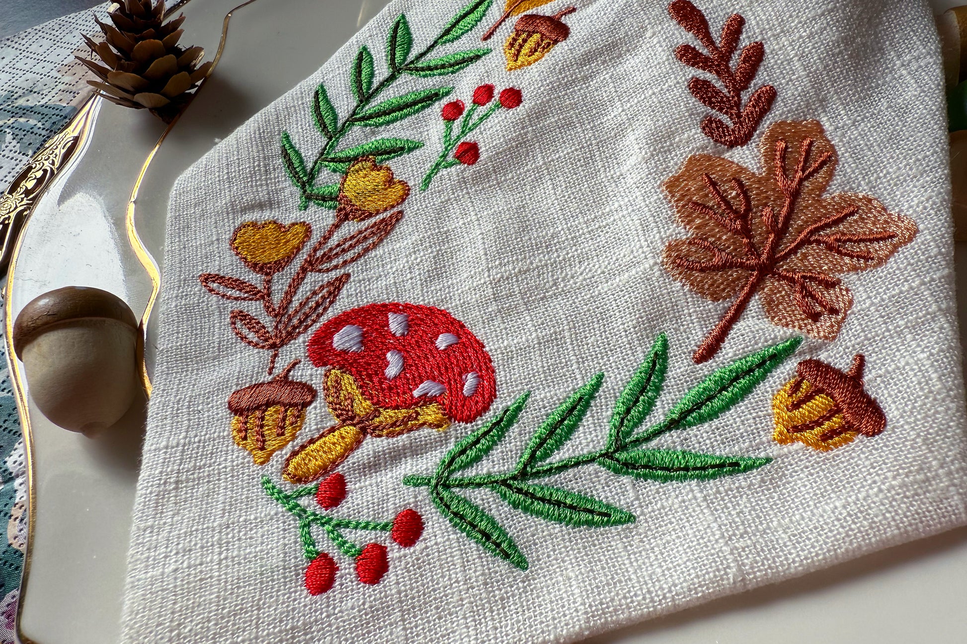 Embroidered fabric with autumn-themed design featuring leaves, acorns, and berries on a white background.