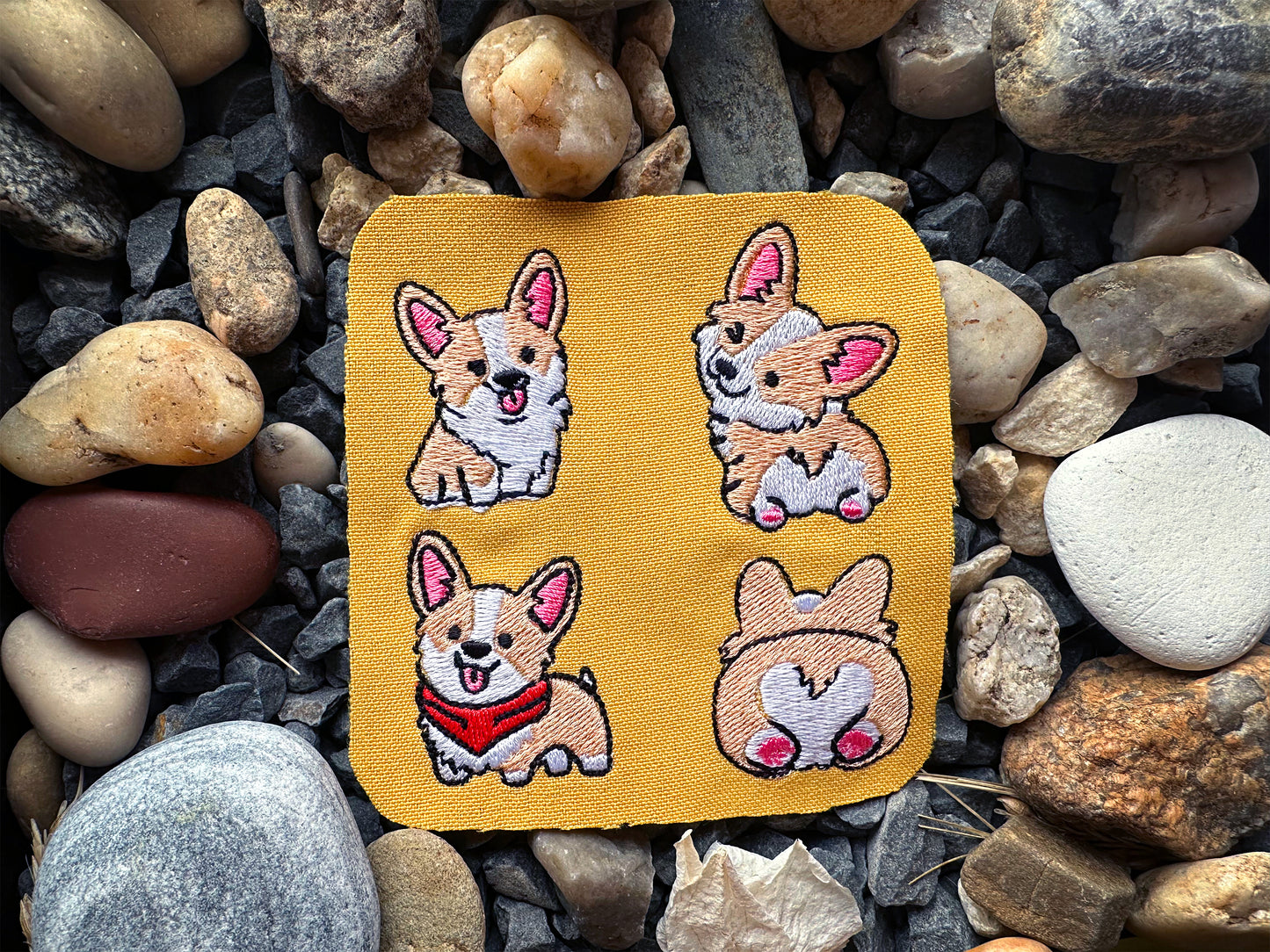 Four embroidered dog patches on a yellow card, placed on a bed of rocks.