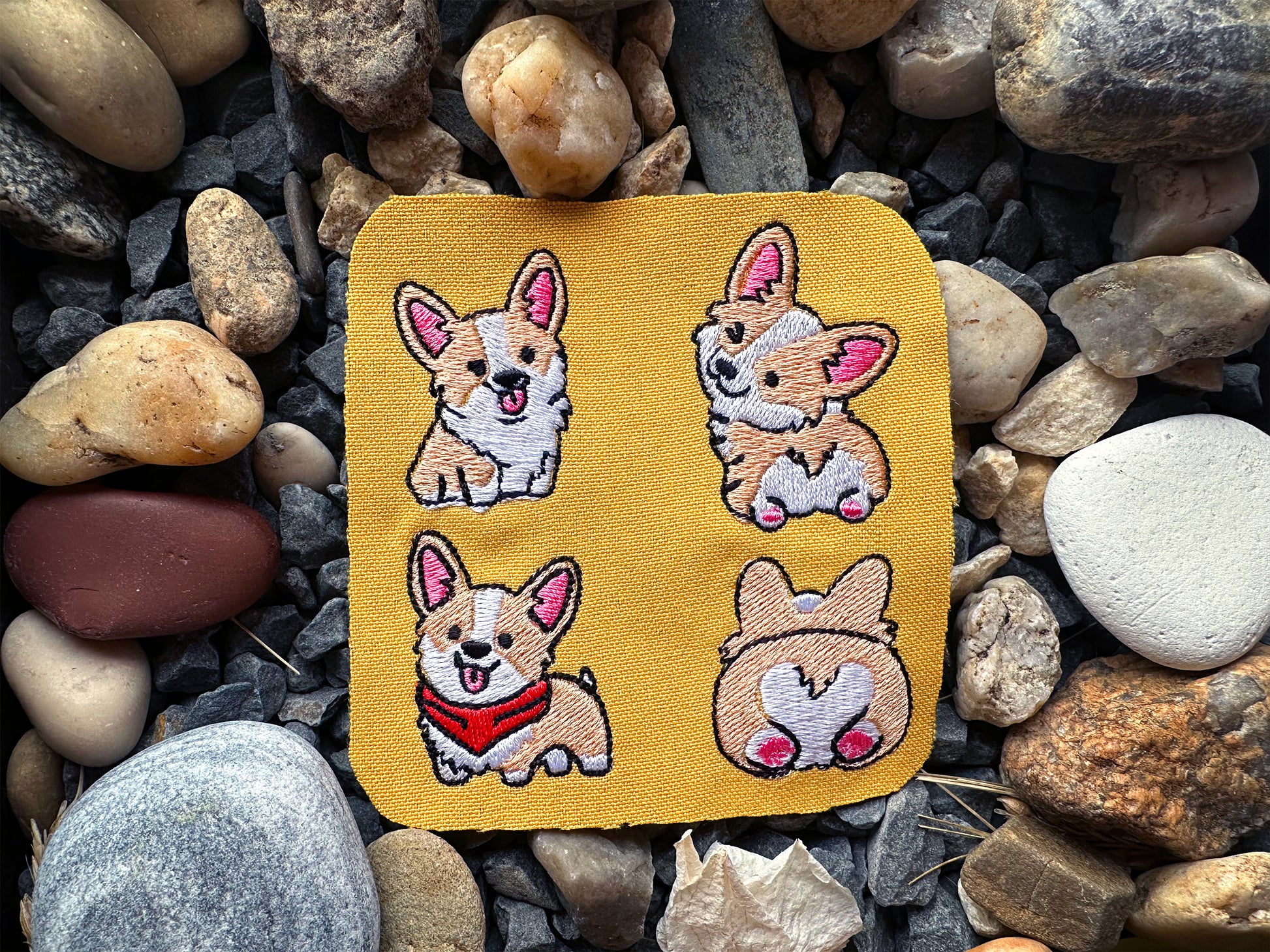 Four embroidered dog patches on a yellow card, placed on a bed of rocks.