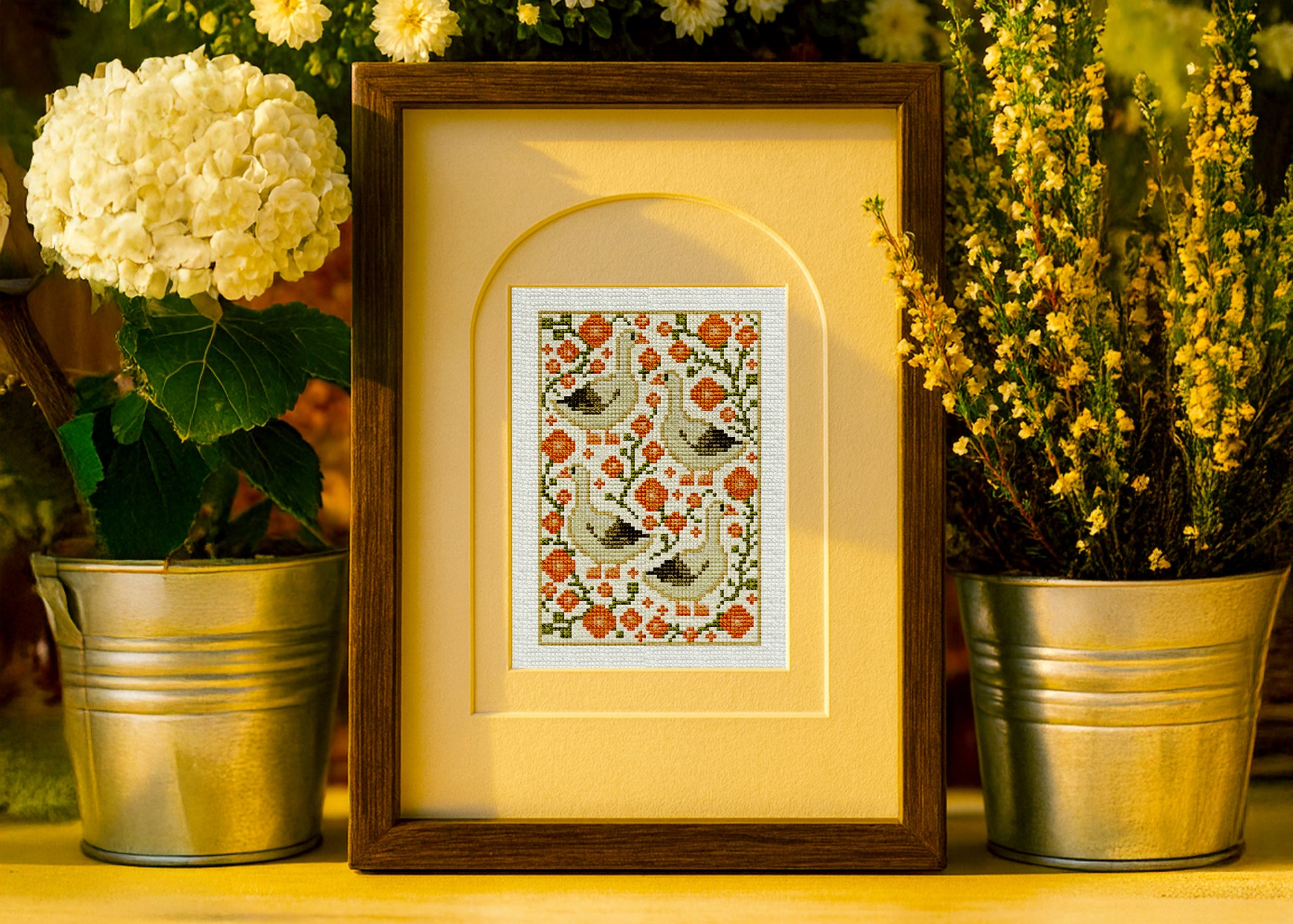 Framed cross-stitched artwork with floral and bird pattern, surrounded by potted plants.