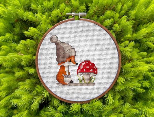 Cross stitch hoop with a little fox in a hat looking at a red mushroom